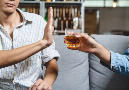 Eine Person in einem karierten Hemd hebt die Hand, um ein alkoholisches Getränk in einem Glas abzulehnen, das von einer anderen Person angeboten wird. Im Hintergrund sind eine Regalwand mit Flaschen und eine gemütliche Raumgestaltung zu sehen.