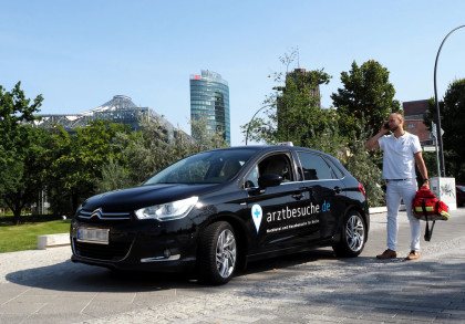 Ein schwarzes Auto mit der Aufschrift "arztbesuche.de" steht am Straßenrand. Daneben steht ein Mann in weißer Kleidung und hält eine rote Tasche sowie ein Mobiltelefon. Im Hintergrund ist eine moderne städtische Umgebung mit Bäumen und Gebäuden zu sehen.