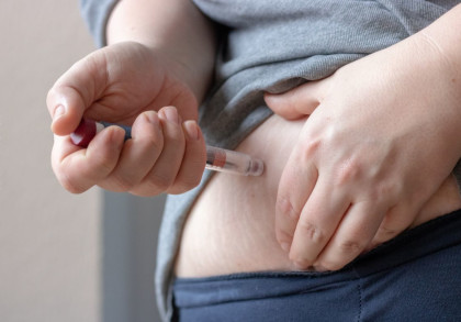 Eine Hand hält eine Spritze und injiziert eine Flüssigkeit in den Bauchbereich einer Person. Die andere Hand zieht die Haut am Bauch leicht zusammen. Die Person trägt ein graues Oberteil und eine dunkelblaue Hose.