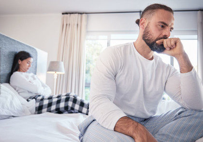 Ein Mann und eine Frau sitzen auf dem Bett und blicken in entgegengesetzte Richtungen. Der Mann im Vordergrund stützt nachdenklich seinen Kopf auf seine Hand, während die Frau im Hintergrund mit verschränkten Armen und gesenktem Kopf sitzt.