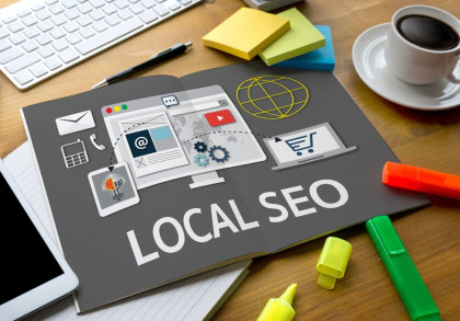 Schreibtisch mit einer Tastatur, einer Tasse Kaffee & Bürobedarf. Im Mittelpunkt steht ein aufgeschlagenes Magazin mit der Aufschrift "LOCAL SEO" und Grafiken zu E-Mail, Onlineshopping, Suchmaschinenoptimierung und Werbung. Notizblöcke und Marker daneben.