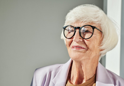Ältere Frau mit weißem Haar und Brille, schaut nach rechts und lächelt leicht. Sie trägt einen hellen Blazer und steht vor einem neutralen Hintergrund.