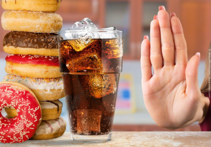Ein Glas Cola mit Eiswürfeln steht neben einem Stapel bunter Donuts. Eine Hand mit ausgestreckter Handfläche signalisiert "Stopp" oder Ablehnung vor den Donuts und dem Getränk.