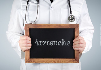 Ein Arzt im weißen Kittel hält eine Tafel mit der Aufschrift "Arztsuche". Um den Hals trägt er ein Stethoskop.