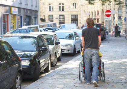 Ein Mann schiebt einen Rollstuhl über einen Bürgersteig auf einer belebten Straße, die von geparkten Autos gesäumt ist. Im Hintergrund sind Geschäfte und mehrstöckige Gebäude zu sehen.