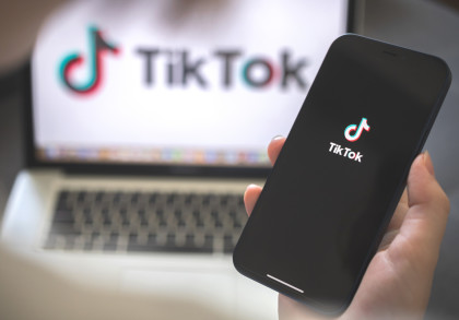  Gemini hat gesagt Eine Hand hält ein schwarzes Smartphone mit dem TikTok-Logo im Vordergrund. Im unscharfen Hintergrund ist ein Laptop-Bildschirm zu sehen, auf dem ebenfalls das TikTok-Logo und der Schriftzug der App abgebildet sind.