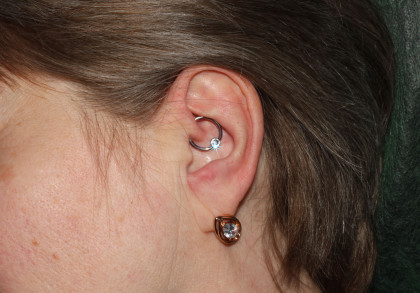 Die Seite eines Kopfes mit Haaren, sowie Teile des Gesichts und Halses. Ein Ohr mit einem Ohrring und einem so genannten Daith Piercing, das im inneren Knorpel des Ohres sitzt.