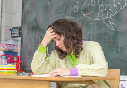 Eine Lehrerin sitzt an ihrem Pult vor der Tafel in einem Klassenzimmer. Sie fasst sich mit der rechten Hand an die Stirn und sieht erschöpft  aus.