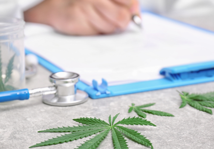 Nahaufnahme von Cannabisblättern, einem Stethoskop und einem Arzt, der im Hintergrund auf einem Klemmbrett schreibt – Symbolbild für medizinisches Cannabis und ärztliche Verschreibung.