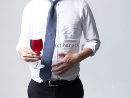 Ein Mann in einem weißen Hemd und einer blauen Krawatte hält ein Glas Rotwein in der Hand und berührt seinen Bauch mit der anderen Hand.