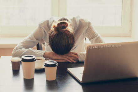 Eine Person mit einem Dutt liegt mit dem Kopf auf verschränkten Armen auf einem Tisch. Vor ihr stehen mehrere Kaffeebecher und ein Laptop. Sie scheint erschöpft oder schläfrig zu sein. Der Raum ist hell erleuchtet durch das Fenster im Hintergrund.