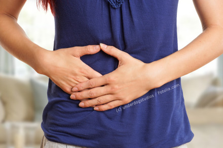 Eine Person in einem blauen Oberteil hält sich ihren Bauch mit beiden Händen, was auf Bauchschmerzen oder Unwohlsein hindeutet.