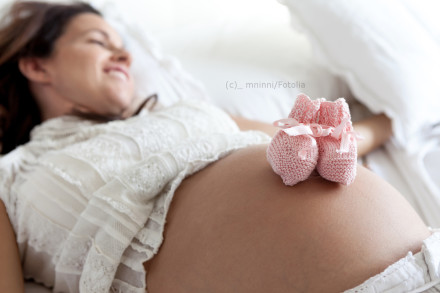 Ein Foto einer lächelnden schwangeren Frau in weißer Spitze, liegend auf einem Bett. Zwei kleine rosafarbene Babyschühchen liegen auf ihrem entblößten Babybauch.