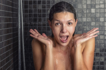 Eine Frau steht unter der Dusche mit nassen Haaren und Wasser, das über ihr Gesicht läuft. Sie hat einen überraschten Gesichtsausdruck und hält ihre Hände zu beiden Seiten ihres Gesichts hoch. Die Duschwände sind mit dunklen Fliesen bedeckt.