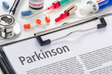 Ein Stethoskop, Pillen, eine Spritze und andere medizinische Utensilien liegen auf einem Schreibtisch. Im Vordergrund befindet sich ein Klemmbrett mit einem Dokument, auf dem das Wort "Parkinson" groß geschrieben steht.