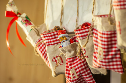 Mehrere kleine Jutesäckchen, mit rot-weiß kariertem Stoff versehen, hängen an Schnüren. In einem der Säckchen steckt eine Schokoladenfigur in Form eines Nikolaus. Ein größeres Geschenk, eingewickelt in Papier mit rotem Band, hängt im Hintergrund.