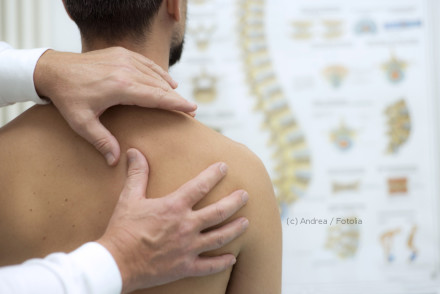 Ein Arzt untersucht den oberen Rücken eines Patienten. Im Hintergrund befindet sich ein Plakat mit anatomischen Darstellungen der Wirbelsäule. Der Patient sitzt mit nacktem Oberkörper vor dem Arzt.