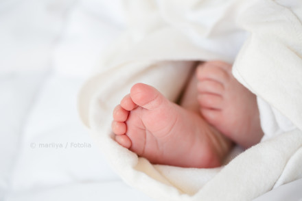 Das Bild zeigt die kleinen Füße eines Babys, eingewickelt in eine weiche, weiße Decke. Die Füße sind nah beieinander und nur teilweise sichtbar. Sie ruhen auf einer weißen Oberfläche und strahlen eine friedliche und zarte Atmosphäre aus.