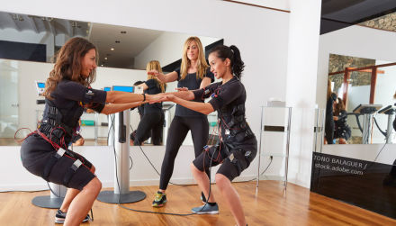Zwei Frauen führen unter der Anleitung einer Trainerin ein EMS-Training (Elektromyostimulationstraining) durch. Beide tragen schwarze Trainingskleidung und sind mit Elektroden und Kabeln verbunden.
