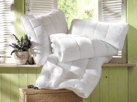 Ein Kissen und eine gefaltete weiße Decke liegen auf einer Fensterbank vor einem Fenster mit weißen Fensterläden. Auf der Fensterbank steht ein dekorativer Topf mit lila Blumen. Die Szene wird von Sonnenlicht erhellt, das durch das Fenster scheint.