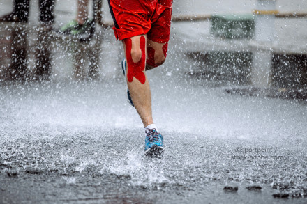 Ein Läufer in roten Shorts und blauen Laufschuhen läuft durch Pfützen und spritzt Wasser bei Regen hoch. Seine Knie sind mit rotem Kinesiologie-Tape beklebt.
