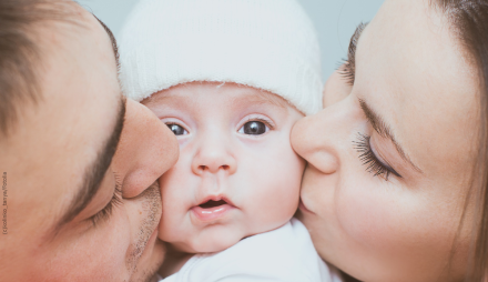 Ein Baby mit einer weißen Mütze wird von zwei Erwachsenen auf ihren Wangen geküsst.