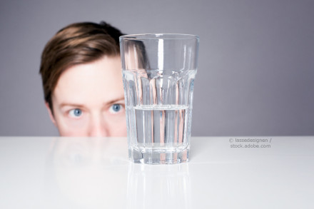 Ein Mann mit blauen Augen schaut neugierig über den Rand eines weißen Tisches. Vor ihm steht ein Glas, das zur Hälfte mit Wasser gefüllt ist. Der Hintergrund ist grau.