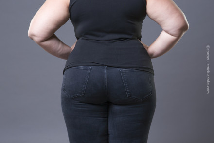 Eine Frau in schwarzem ärmellosem Oberteil und schwarzer Jeanshose, die von hinten fotografiert wird, ihre Hände in die Hüften gestützt.