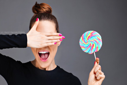Eine Frau mit einem Haarknoten hält einen farbenfrohen Lutscher in der rechten Hand und verdeckt ihr linkes Auge mit der linken Hand. Sie trägt schwarzen Pullover und hat pink lackierte Fingernägel. Der Hintergrund ist grau.