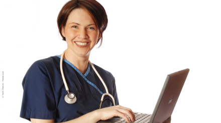 Eine lächelnde Frau in blauer medizinischer Arbeitskleidung, mit einem Stethoskop um den Hals, arbeitet an einem Laptop. Der Hintergrund ist weiß.