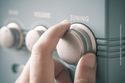 Nahaufnahme einer Hand, die den Abstimmknopf eines Radios dreht. Im Hintergrund sind weitere Bedienknöpfe für Lautstärke und eine unscharfe Markierung zu erkennen.