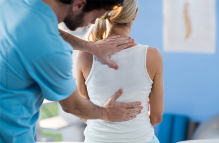 Ein Physiotherapeut in einem blauen T-Shirt massiert den Rücken einer Frau mit blonden Haaren, die ein weißes Tanktop trägt. Die Umgebung deutet auf eine Physiotherapiepraxis hin.