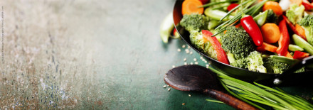 Ein Topf mit frisch geschnittenem Gemüse, inklusive Brokkoli, Paprika, Karotten & Chilis, liegt auf einem rustikalen Tisch. Ein hölzerner Kochlöffel und ein paar grüne lange Blätter liegen daneben. Im Hintergrund ist ein verwaschener, grüner Untergrund.