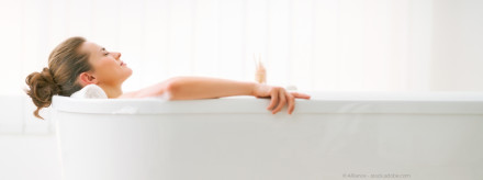 Eine Frau mit hochgestecktem braunen Haar entspannt sich in einer Badewanne und lehnt sich zurück. Sie hat die Augen geschlossen und wirkt ruhig. Im Hintergrund ist verschwommenes weißes Licht zu sehen.