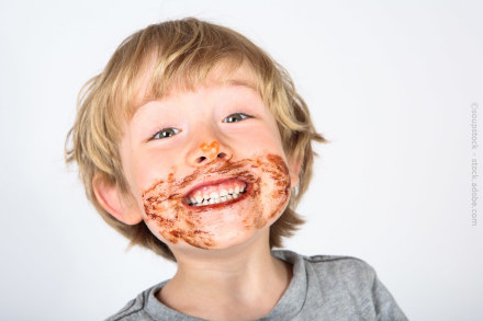 Ein kleiner Junge mit blonden Haaren lächelt breit in die Kamera. Sein Gesicht ist mit Schokoladenflecken verschmiert, besonders um die Lippen und auf der Nase. Er trägt ein graues T-Shirt. Der Hintergrund ist weiß.
