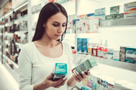 Eine Frau mit langen, dunklen Haaren steht in einer Apotheke und hält zwei verschiedene Medikamentenpackungen in den Händen. Sie trägt ein weißes Oberteil und scheint die Informationen auf den Verpackungen zu lesen.