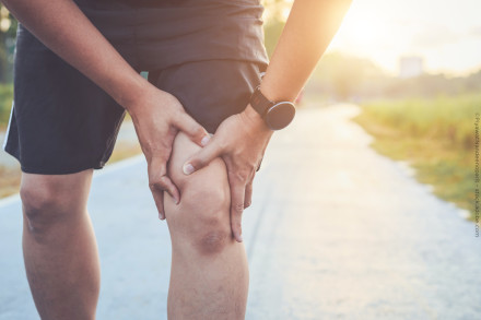 Eine Person, die sich an das Knie hält und Schmerzen hat, während sie auf einem Pfad im Freien steht. Die Person trägt eine Uhr und Shorts, und im Hintergrund ist ein sonniger Tag zu sehen.