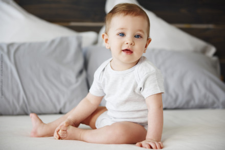 Das Bild zeigt ein Kleinkind, das auf einem Bett sitzt. Das Kind trägt einen hellgrauen Body und lächelt leicht in die Kamera. Im Hintergrund sind weiße und graue Kissen zu sehen.