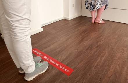 Zwei Personen stehen in einem Raum auf einem Holzboden. Die eine Person trägt weiße Hosen und graue Sneakers, die andere eine gemusterte Kleidung und weiße Sandalen.
