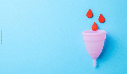 Das Bild zeigt eine rosa Menstruationstasse auf einem hellblauen Hintergrund. Über der Tasse schweben drei rote Tropfen.