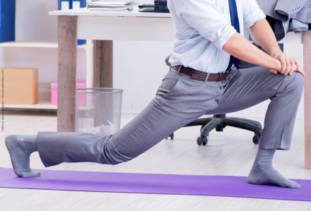 Ein Mann in Businesskleidung macht eine Dehnübung auf einer lila Yogamatte in einem Büro. Im Hintergrund sind ein Tisch mit Büromaterialien, ein Mülleimer und ein Regal zu sehen.