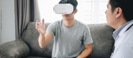 Ein Mann sitzt auf einem Sofa und trägt ein Virtual-Reality-Headset. Er hebt eine Hand, als ob er etwas in der virtuellen Welt berührt. Neben ihm sitzt eine weitere Person und beobachtet ihn. Im Hintergrund sind ein Fenster & ein grauer Vorhang zu sehen.