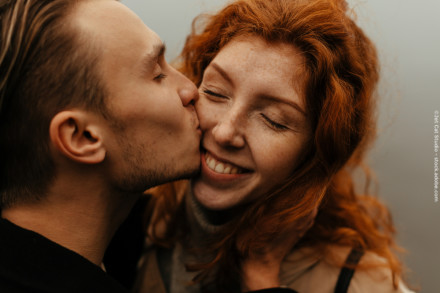 Ein Mann küsst eine lächelnde Frau auf die Wange. Die Frau hat rote Haare und Sommersprossen und ihre Augen sind geschlossen, während sie glücklich lächelt.