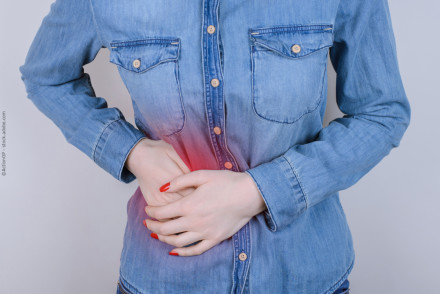 Eine Person in einem Jeanshemd hält sich den schmerzenden unteren Bauchbereich mit beiden Händen. Der Schmerzbereich ist durch eine rote Färbung hervorgehoben.