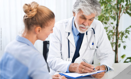 Ein älterer männlicher Arzt mit grauem Haar und Bart sitzt an einem Schreibtisch. Er trägt einen weißen Arztkittel und ein Stethoskop um den Hals. Der Arzt ist in ein Gespräch mit einer jungen Frau vertieft, die auf der linken Seite des Bildes sitzt.