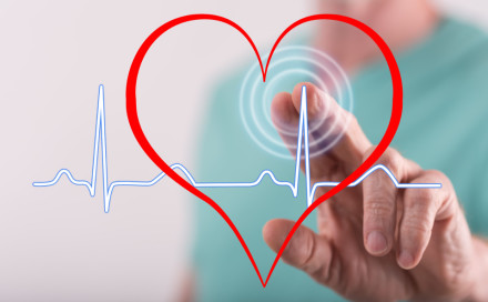 Eine Hand zeigt auf ein großes rotes Herz mit einem digitalen Herzschlag-Muster auf einem transparenten Hintergrund. Im Fokus steht die grafische Darstellung eines Herzfrequenzverlaufs als Symbol für Gesundheit und Medizin.