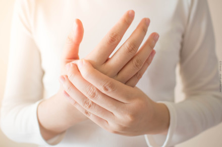 Nahaufnahme einer Person, die ihre schmerzende Hand hält und eine weiße Kleidung trägt.