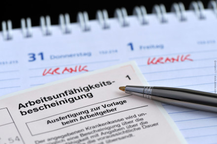 Eine Nahaufnahme zeigt eine Arbeitsunfähigkeitsbescheinigung und einen Kugelschreiber, die auf einem Kalender liegen. Im Kalender sind zwei Tage, Donnerstag und Freitag, mit der handschriftlichen Notiz „krank“ markiert.