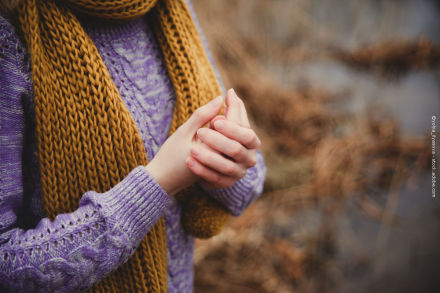 Nahaufnahme einer Person, die einen lila Strickpullover und einen senfgelben Strickschal trägt. Die Person hält ihre Hände vor dem Körper. Der Hintergrund ist unscharf und zeigt eine herbstliche Landschaft.