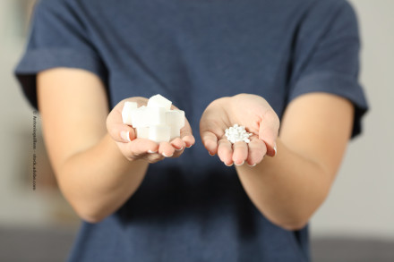 Eine Person in einem dunkelblauen T-Shirt hält in einer Hand mehrere weiße Zuckerstücke und in der anderen Hand weiße, kleine Tabletten.